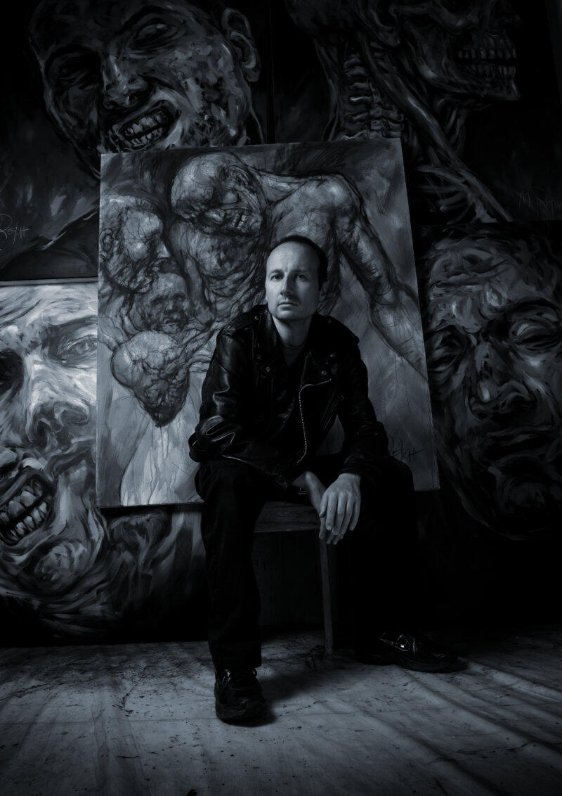 Black and white photo of artist painter Marcel Reich in front of his large-scale paintings, featuring a visceral, dark surrealism with a dystopic edge.